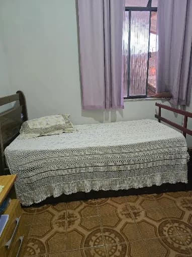 Quarto com cama de solteiro coberta por colcha de crochê, cortina clara e janela com luz natural