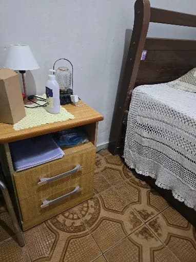 Criado-mudo de madeira ao lado da cama, com abajur e objetos pessoais do morador